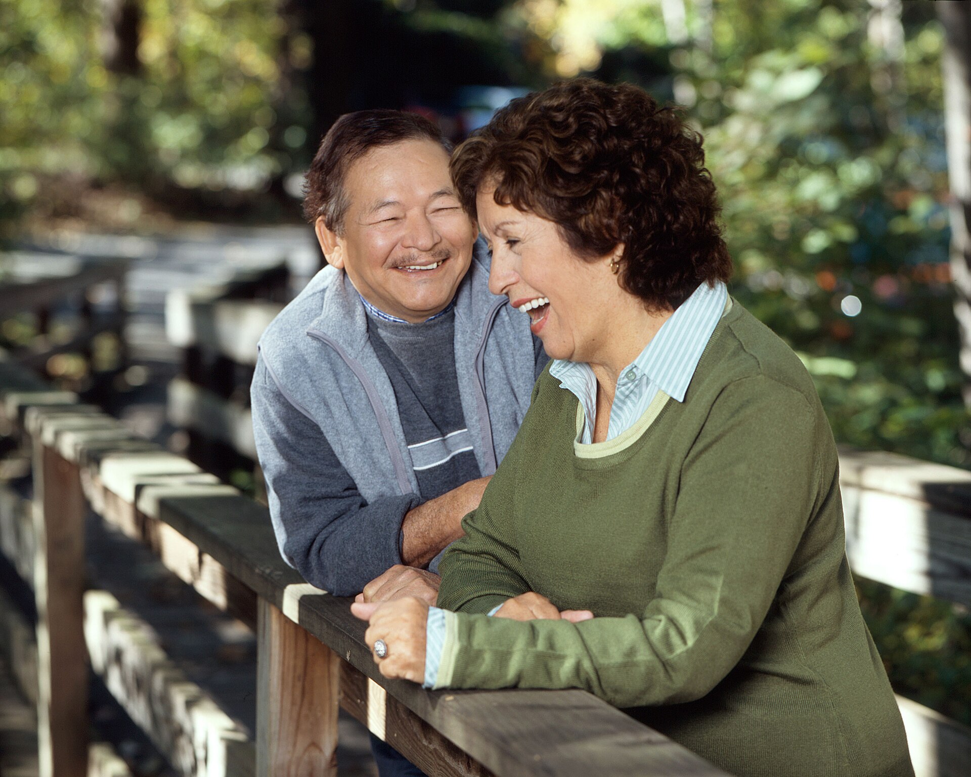 A man and a woman laughing together while leaning on a wooden railing outdoors.