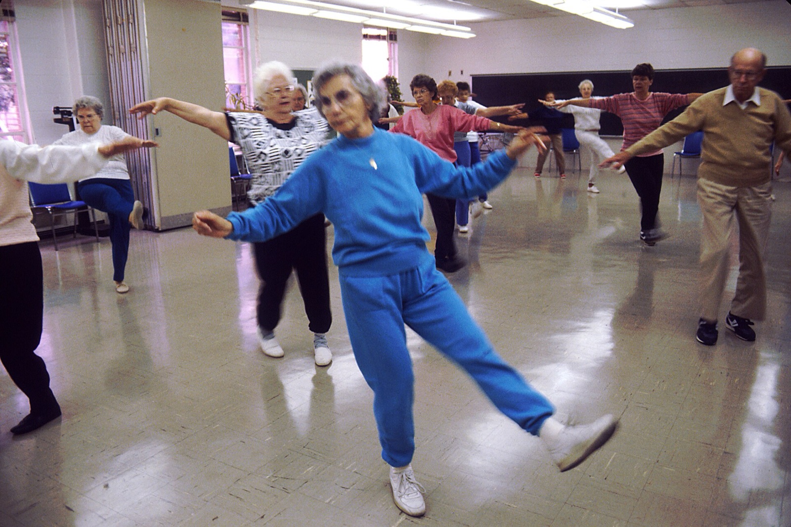 A group of older adults doing a stretching and balance exercise class in a bright hall.