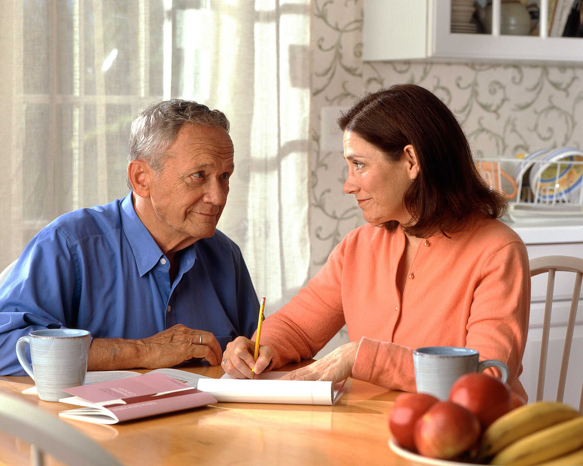 A woman and a man talking over notes and a pencil at a sunny kitchen table.